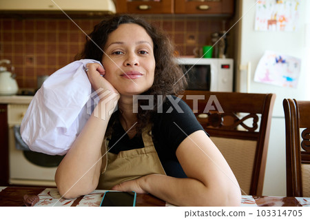 Beautiful curly haired multi ethnic woman housewife, baker confectioner in chef's apron, sitting at table in home kitchen, smiling looking confidently at camera. Culinary. Cookery. People and hobby 103314705