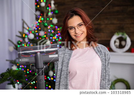 An attractive girl holds a vacuum cleaner in her hand against the background of a Christmas tree. An attractive girl holds a vacuum cleaner in her hand against the background of a Christmas tree. 103318985