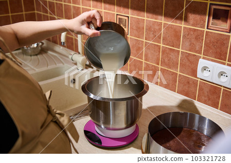 Close-up female pastry chef pouring fresh cream into a stainless steel bowl of food processor with planetary mixer, weighing ingredients on kitchen scale, preparing whipped cream for chocolate cake 103321728