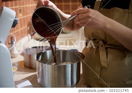Close-up pastry chef pouring melted chocolate into a metal bowl of planetary mixer, mixing it with cream for preparing delicious chocolate cream or with whipped egg whites for making sponge biscuit 103321745