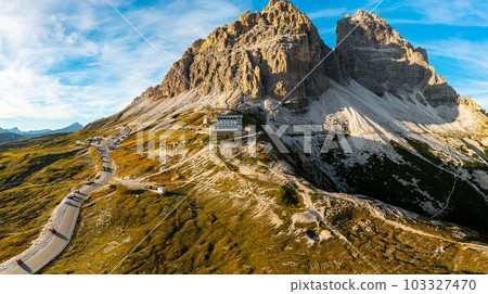 Aerial View of Tre Cime di Lavaredo, Serpentine Road Embraces Majestic Rocky Mountain Amidst Scenic Mountain Ranges Bathed in Sunset Glowx. Aerial View of Tre Cime di Lavaredo, Serpentine Road Embraces Majestic Rocky Mountain Amidst Scenic Mountain Ranges Bathed in Sunset Glowx. 103327470