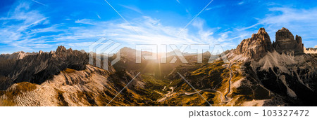 Curved road on rocky highland under blue sky with white clouds. Bare mountain landscape of Three Peaks of Lavaredo at sunset aerial view Curved road on rocky highland under blue sky with white clouds. Bare mountain landscape of Three Peaks of Lavaredo at sunset aerial view 103327472