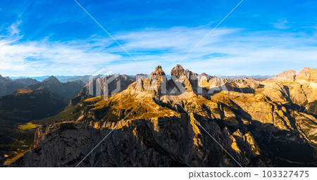 Mountain ranges of Alps under blue sky with white clouds. Rocky peaks above wide shaded canyon. Three peaks of Lavaredo at sunset aerial view Mountain ranges of Alps under blue sky with white clouds. Rocky peaks above wide shaded canyon. Three peaks of Lavaredo at sunset aerial view 103327475