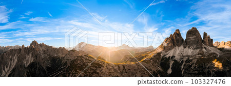 Sun sets on horizon above rocky mountain range. Sand-covered slopes of foothills of Tre Cime di Lavaredo silhouette at sunset aerial view in back lit Sun sets on horizon above rocky mountain range. Sand-covered slopes of foothills of Tre Cime di Lavaredo silhouette at sunset aerial view in back lit 103327476
