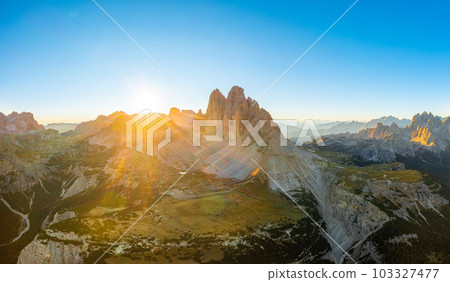 Sun rises from behind peak of rocky mountain silhouette. Landscape of Dolomite Alps under clear sky. Tre Cime di Lavaredo at sunrise aerial view in back lit Sun rises from behind peak of rocky mountain silhouette. Landscape of Dolomite Alps under clear sky. Tre Cime di Lavaredo at sunrise aerial view in back lit 103327477