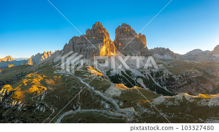 Sharp faces of high mountains at sunrise. Illuminated by sun rays rocky peaks of Tre Cime di Lavaredo under clear sky aerial view in back lit Sharp faces of high mountains at sunrise. Illuminated by sun rays rocky peaks of Tre Cime di Lavaredo under clear sky aerial view in back lit 103327480