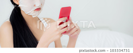 Beautiful young asian woman with sheet facial mask sitting watching smartphone on bed. 103334731
