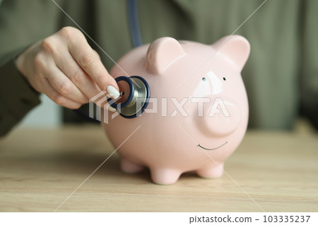 Female person checks piggy bank condition with stethoscope Female person checks piggy bank condition with stethoscope 103335237