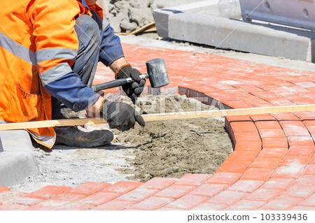 A worker using a rubber mallet and a wooden level lays orange paving slabs evenly on a sunny summer day. 103339436