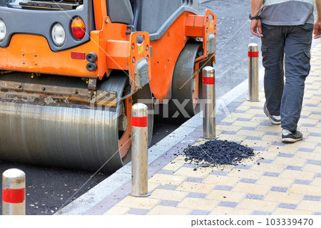 The metal wheels of the agile road roller compact the fresh asphalt on the stretch of road along the sidewalk. The metal wheels of the agile road roller compact the fresh asphalt on the stretch of road along the sidewalk. 103339470