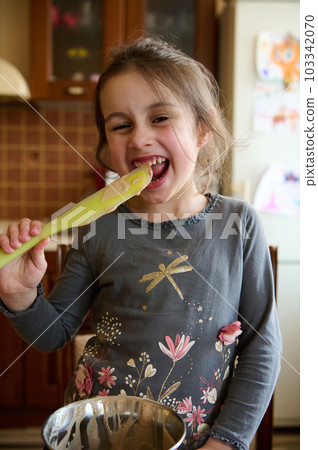 Cute little girl helps her mom in kitchen to prepare cream for the cake, quietly licks a spoon with melted delicious confectionery chocolate, mischievously smiling and making faces looking at camera Cute little girl helps her mom in kitchen to prepare cream for the cake, quietly licks a spoon with melted delicious confectionery chocolate, mischievously smiling and making faces looking at camera 103342070