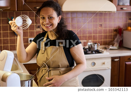 Beautiful multi ethnic pregnant woman, charming pretty housewife, smiling looking at camera showing a planetary mixer whisk with whipped cream, preparing sweet mousse dessert in her cozy home kitchen Beautiful multi ethnic pregnant woman, charming pretty housewife, smiling looking at camera showing a planetary mixer whisk with whipped cream, preparing sweet mousse dessert in her cozy home kitchen 103342113
