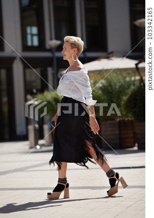 Confidence and sophistication mature woman walks city streets in fashionable ensemble chic white shirt and black fringed skirt with high heeled sandals and socks, fashion icon of style 103342165