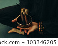 Top view of two hands of a man holding a singing bowl with sticks in front of him. Listen to the healing vibrations of Tibetan bowls. Humility and calm. Rosary on the hand. Close-up 103346205