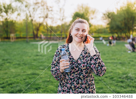 A portrait of a smiling beautiful woman talking on phone on nature background. Happy woman in dress is using a smartphone in park outdoors, summer time. Traveler 103346488