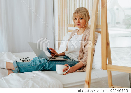 Senior mature woman sitting on bed with coffee tea mug and debit credit card while using her laptop for online shopping investments insurance Senior mature woman sitting on bed with coffee tea mug and debit credit card while using her laptop for online shopping investments insurance 103346733