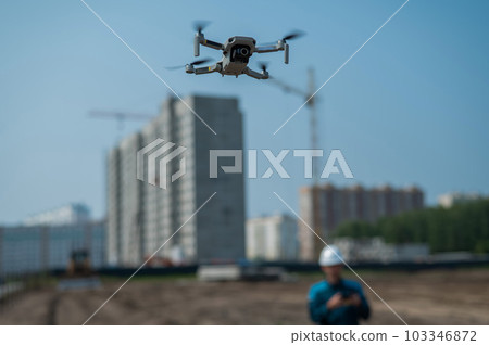 A man in a helmet and overalls controls a drone at a construction site. The builder carries out technical oversight. 103346872