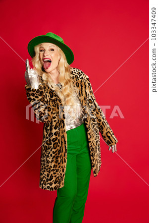 Portrait of beautiful senior lady, woman in stylish clothes, coat and green hat posing against red studio background. Cheeky mood 103347409