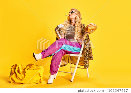 Portrait of elegant senior woman in sportswear, heeled boots and animal print coat sitting and smoking against yellow studio background 103347417