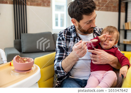 Father feeding crying capricious infant daughter sitting at home. 103347642