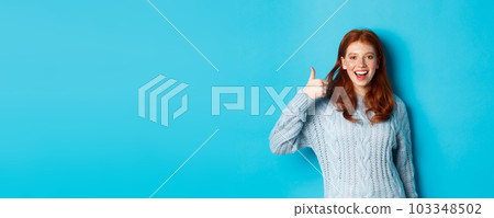 Cheerful redhead girl in sweater, showing thumb up in approval, like and praise product, standing over blue background Cheerful redhead girl in sweater, showing thumb up in approval, like and praise product, standing over blue background 103348502
