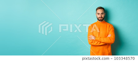 Confident young man looking determined, cross arms on chest, wearing orange winter sweater, standing against turquoise studio background Confident young man looking determined, cross arms on chest, wearing orange winter sweater, standing against turquoise studio background 103348570