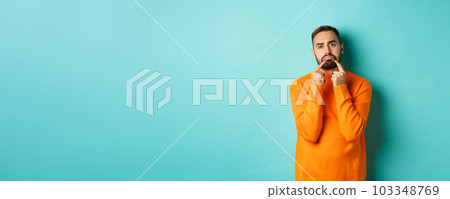 Image of gloomy bearded man, making sad face and frowning, standing upset in orange sweater against turquoise background 103348769