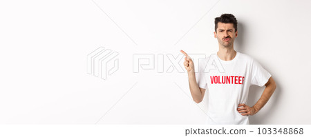 Skeptical and hesitant male volunteer in t-shirt grimacing doubtful, pointing finger left at promo offer, white background 103348868
