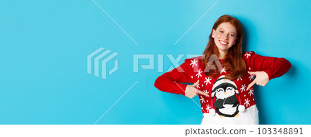 Winter holidays and Christmas Eve concept. Pretty redhead girl pointing fingers at cute xmas sweater with penguin, standing over blue background 103348891