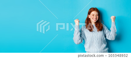 Satisfied redhead girl achieve goal and celebrating, making fist pump gesture and smiling with rejoice, triumphing of win, standing against blue background Satisfied redhead girl achieve goal and celebrating, making fist pump gesture and smiling with rejoice, triumphing of win, standing against blue background 103348932