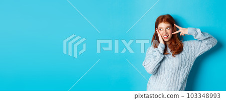 Happy redhead girl smiling, showing peace sign and looking left at promo, standing in sweater against blue background Happy redhead girl smiling, showing peace sign and looking left at promo, standing in sweater against blue background 103348993