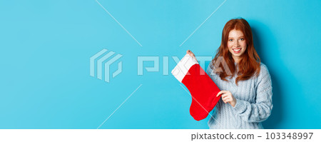 Winter holidays and gifts concept. Cute redhead girl in sweater showing Christmas stocking and smiling, celebrating New Year, standing over blue background Winter holidays and gifts concept. Cute redhead girl in sweater showing Christmas stocking and smiling, celebrating New Year, standing over blue background 103348997