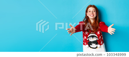 Winter holidays and Christmas Eve concept. Beautiful redhead girl in xmas sweater reaching for hug, extend hands for cuddles and smiling, standing in sweater against blue background Winter holidays and Christmas Eve concept. Beautiful redhead girl in xmas sweater reaching for hug, extend hands for cuddles and smiling, standing in sweater against blue background 103348999