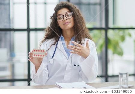 Young pharmacist woman holding pills while sitting at the table in the office Young pharmacist woman holding pills while sitting at the table in the office 103349090