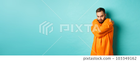 Timid man feeling offended and defensive, hugging himself and looking suspicious at camera, standing over light blue background Timid man feeling offended and defensive, hugging himself and looking suspicious at camera, standing over light blue background 103349162