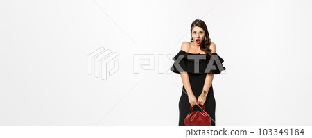 Beauty and fashion concept. Full length if silly young woman pouting and looking surprised, holding purse, wearing heels and black dress, white background Beauty and fashion concept. Full length if silly young woman pouting and looking surprised, holding purse, wearing heels and black dress, white background 103349184