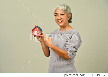 Beautiful mature woman hold small house model isolated on beige background. Real estate, building, property and insurance concept 103349187