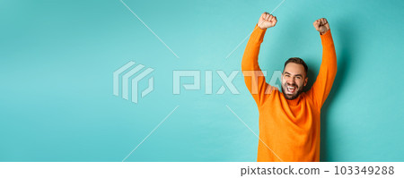 Waist-up shot of happy guy triumphing, raising hands up and rejoicing of winning, celebrating victory, standing against turquoise background Waist-up shot of happy guy triumphing, raising hands up and rejoicing of winning, celebrating victory, standing against turquoise background 103349288