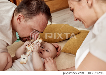 A cute joyful baby and his parents lie on the floor in a bright cozy room with toys. Mom and dad hug and kiss their baby. Happy parenthood and childhood. 103349296