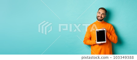 Technology. Handsome adult man showing digital tablet screen, smiling and looking at upper left corner thoughtful, imaging or daydreaming, blue background Technology. Handsome adult man showing digital tablet screen, smiling and looking at upper left corner thoughtful, imaging or daydreaming, blue background 103349388