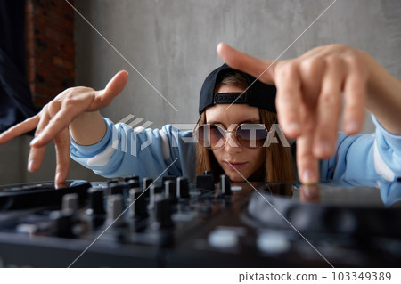 A young pretty long-haired DJ girl in a blue sweater and black baseball cap poses with a black DJ mixing console and mixes music tracks. Close-up studio shot, gray background. A young pretty long-haired DJ girl in a blue sweater and black baseball cap poses with a black DJ mixing console and mixes music tracks. Close-up studio shot, gray background. 103349389