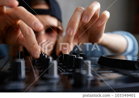 A young pretty long-haired DJ girl in a blue sweater and black baseball cap poses with a black DJ mixing console and mixes music tracks. Close-up studio shot, gray background. 103349401