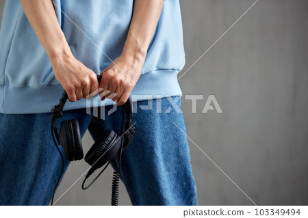 A young pretty DJ girl in a blue sweater and jeans holds black headphones in her hands. Close-up studio shot, gray background. 103349494