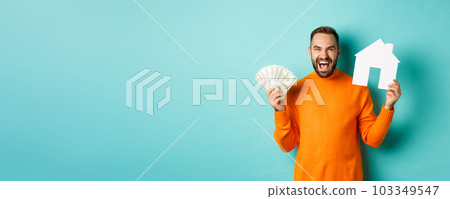 Real estate concept and mortgage concept. Ecstatic caucasian man holding money dollars and house maket, shouting for joy, standing over blue background 103349547