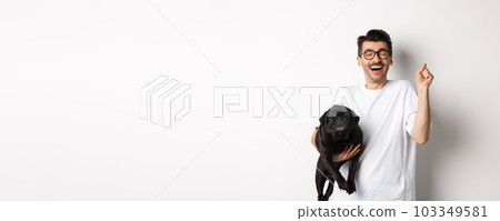 Cheerful handsome man with dog rejoicing, celebrating victory. Happy guy carry cute black pug and looking upbeat, adopting pet, standing over white background Cheerful handsome man with dog rejoicing, celebrating victory. Happy guy carry cute black pug and looking upbeat, adopting pet, standing over white background 103349581