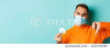 Real estate and coronavirus pandemic concept. Close-up of disappointed man in face mask, showing small paper house and thumb-down, turquoise background 103349584