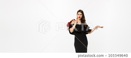 Beauty and fashion concept. Full length of arrogant and sassy woman in black dress, holding purse on shoulder and stroll forward at camera, posing over white background 103349640