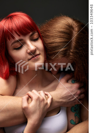 A young attractive woman with bright ruby hair in the arms of a semi-naked man with long hair. The lover hugs the girl from behind. Studio shot, dark background. A young attractive woman with bright ruby hair in the arms of a semi-naked man with long hair. The lover hugs the girl from behind. Studio shot, dark background. 103349648