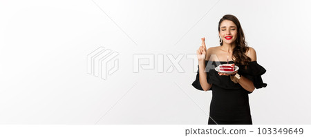 Celebration and party concept. Hopeful and dreamy woman making wish on birthday cake, cross fingers and smiling happy, standing over white background Celebration and party concept. Hopeful and dreamy woman making wish on birthday cake, cross fingers and smiling happy, standing over white background 103349649