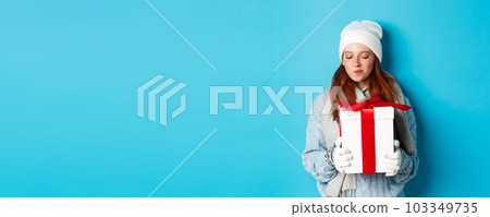 Winter holidays and Christmas sales concept. Intrigued redhead girl holding present, curiously staring at box with gift, trying guess what inside, standing over blue background 103349735